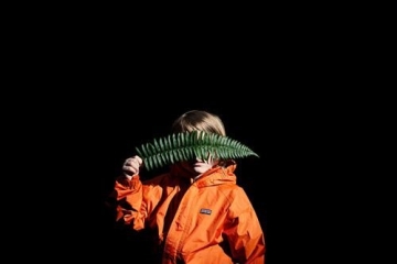photograph of young child in orange jacket holding a green fern against a black background