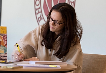 English student writing at table with RIC seal in background on wall