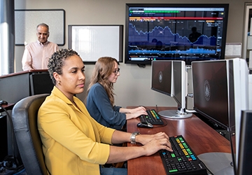 Two students work in finance lab with mounted screen showing financials and professor in background