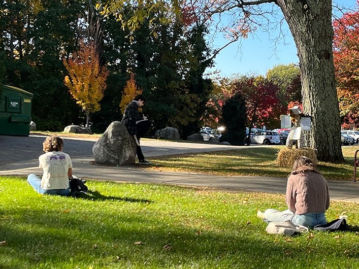 Backs of two people sitting on campus grass, writing in the sunlight