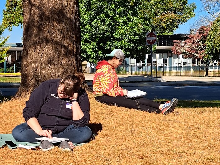 Two people writing under a tree, sitting on pine needles, on campus