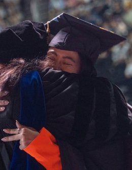 Graduate in cap and gown at ceremony in heartfelt embrace with faculty member